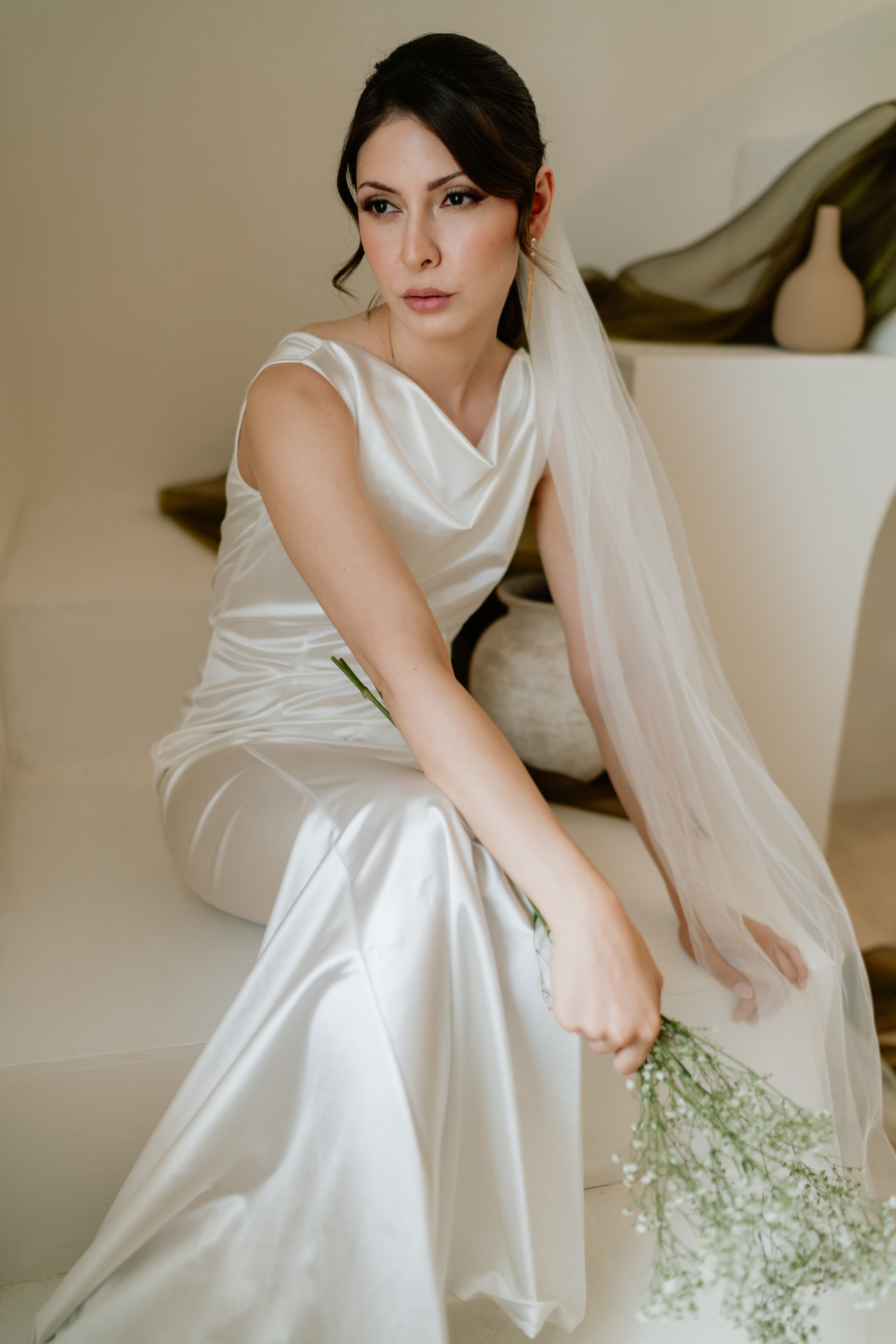 Bride seated in a satin gown and long veil holding a small bouquet.