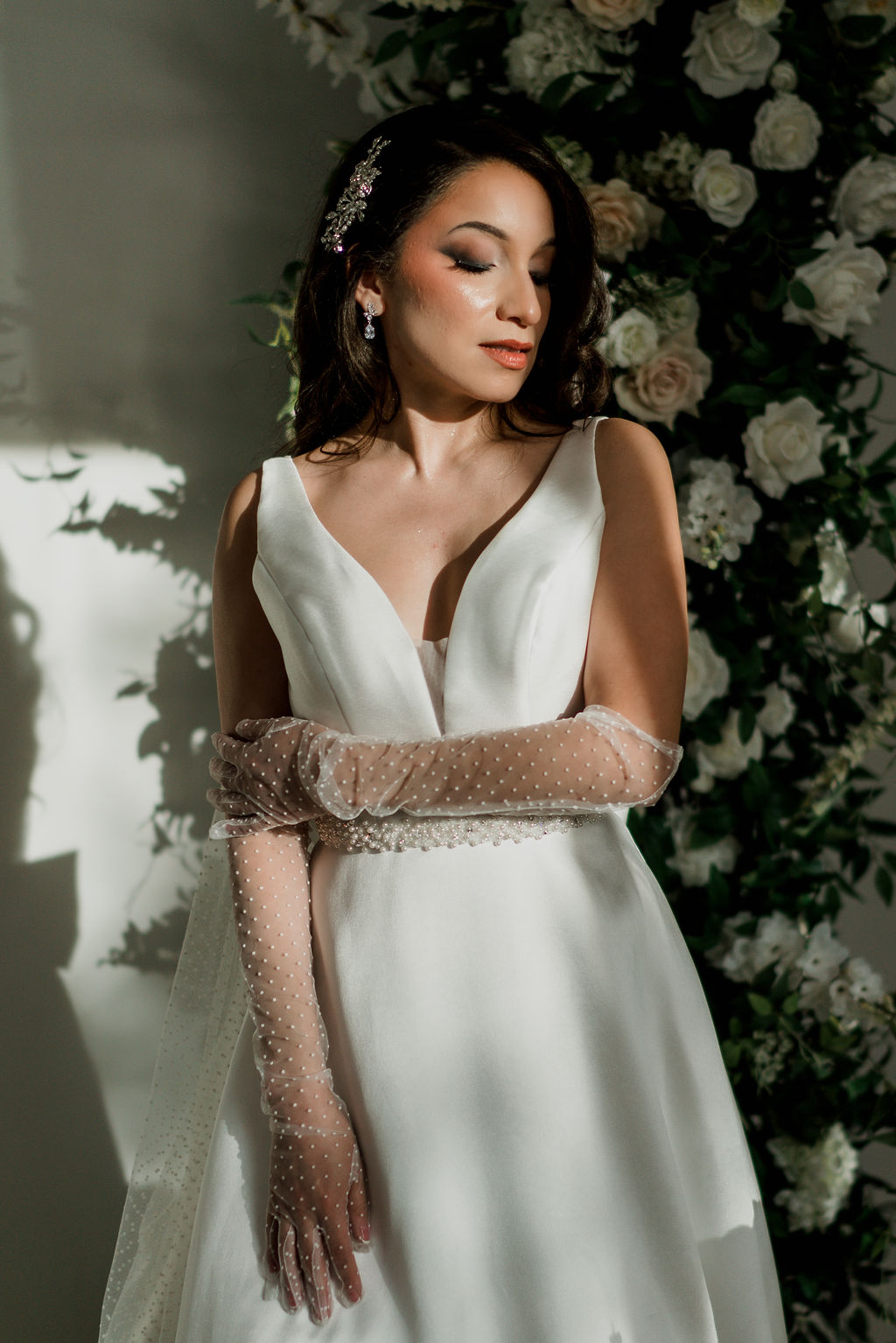 Bride with a sleek side-swept look, crystal hair piece, and sheer dotted gloves in front of a wall of white roses.