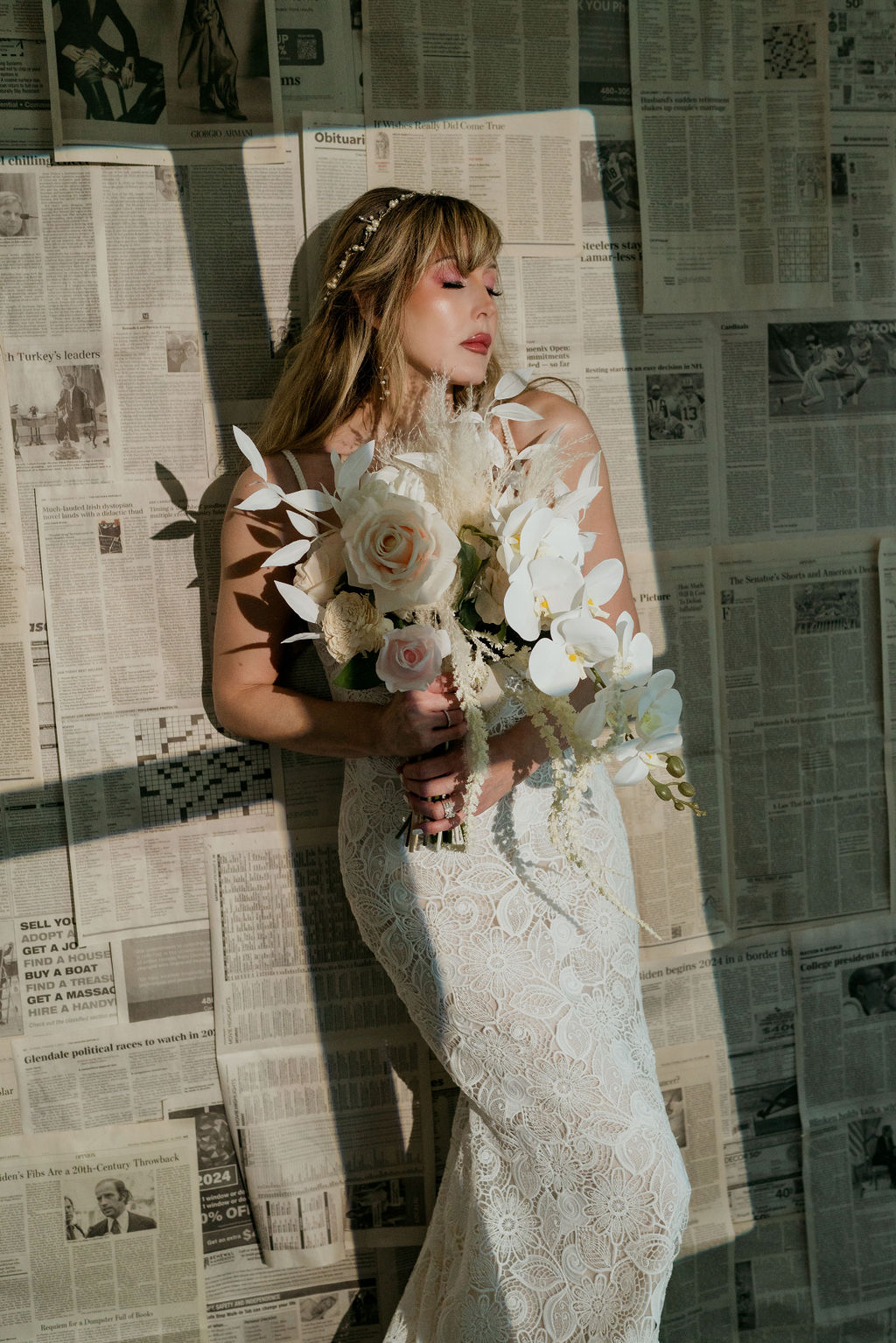 Blonde bride with bangs, a pearl hair vine, and a white bouquet, leaning against a wall of newspapers in the sun.