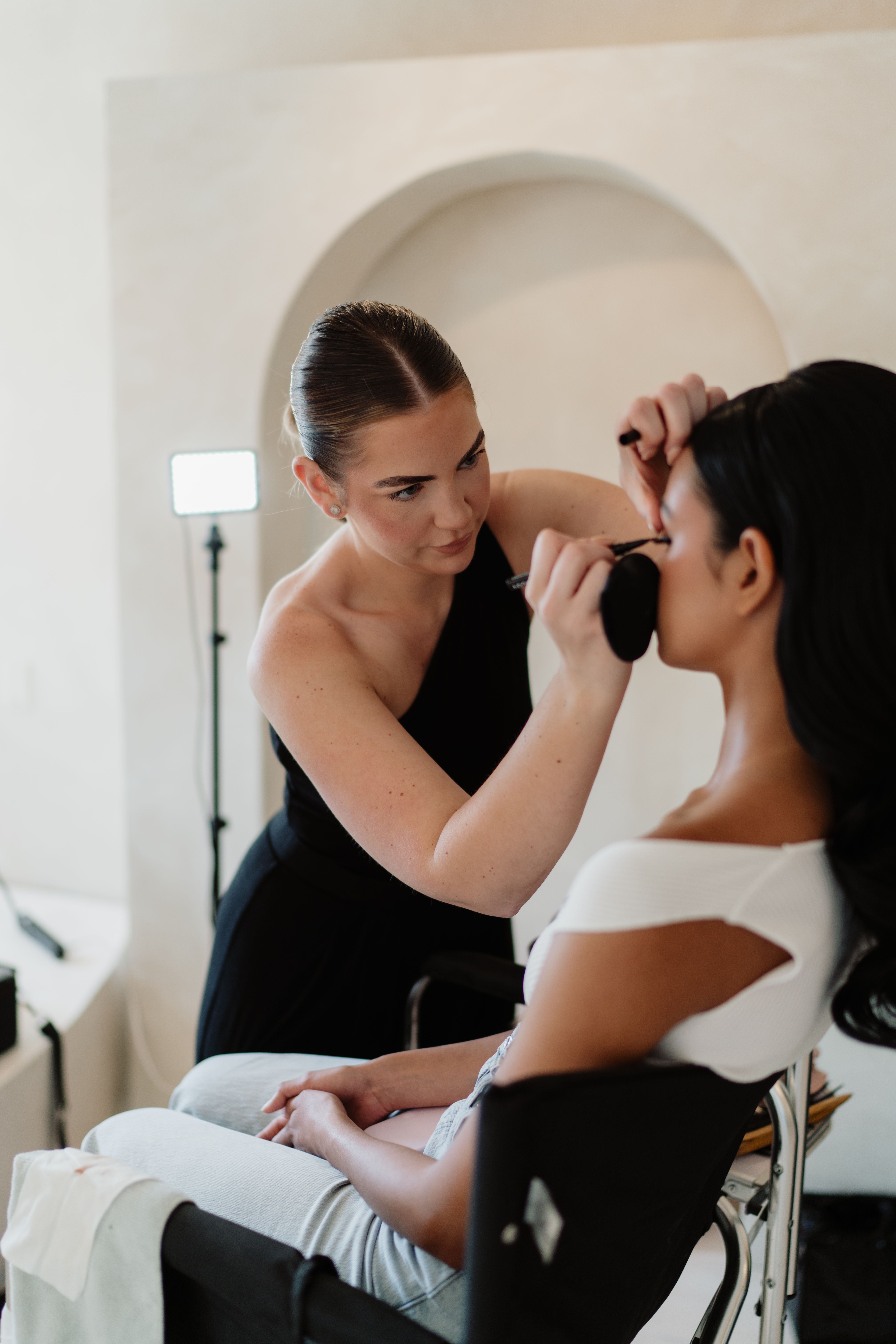 A professional makeup artist in a black top carefully applies eyeliner to a seated client in a bright, modern studio.