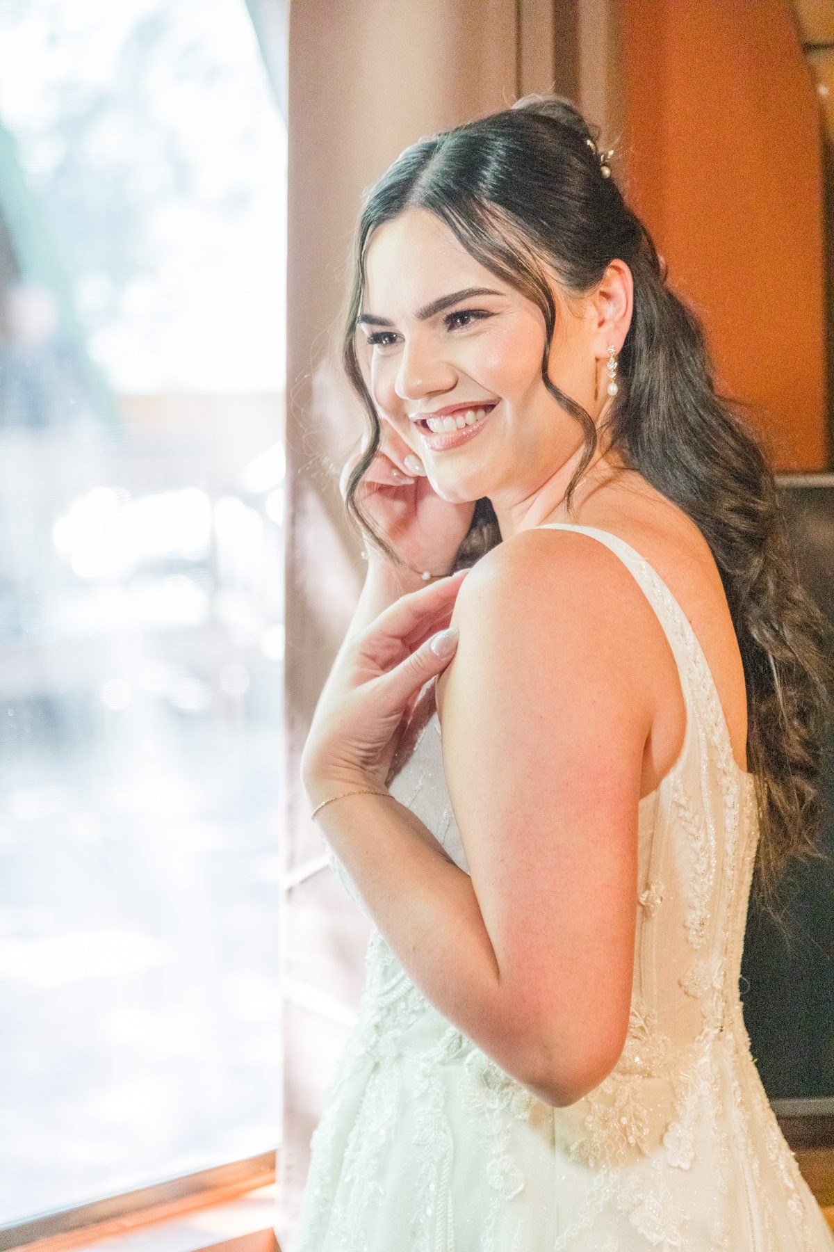 Bianca smiling by a window on her wedding day, with soft glam and a half-up hairstyle