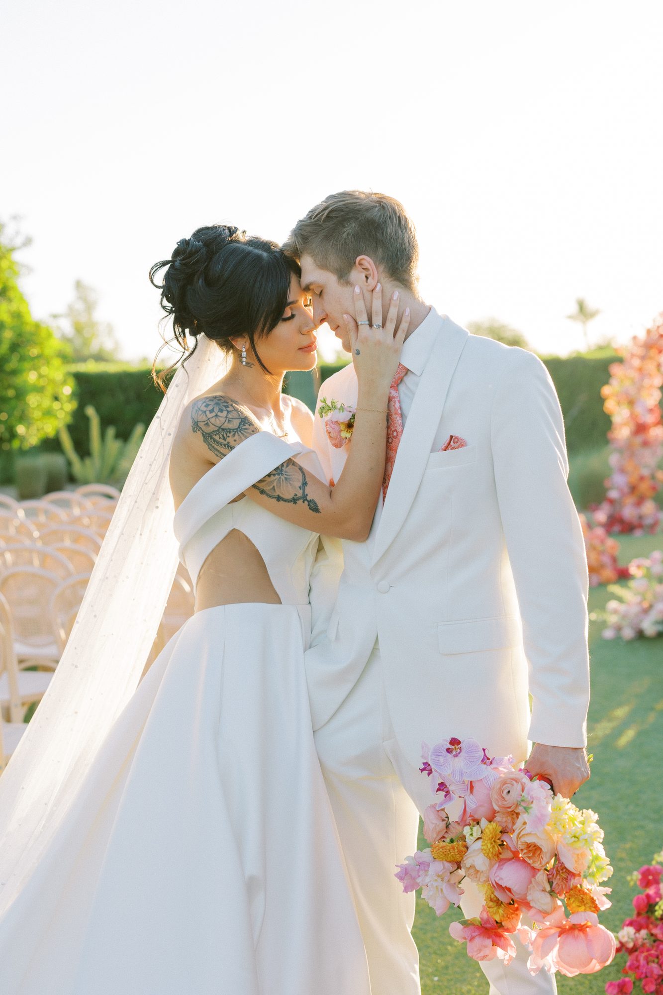 Bride with a sleek updo and soft veil leaning into her groom in a white suit, holding a bright bouquet in a garden.