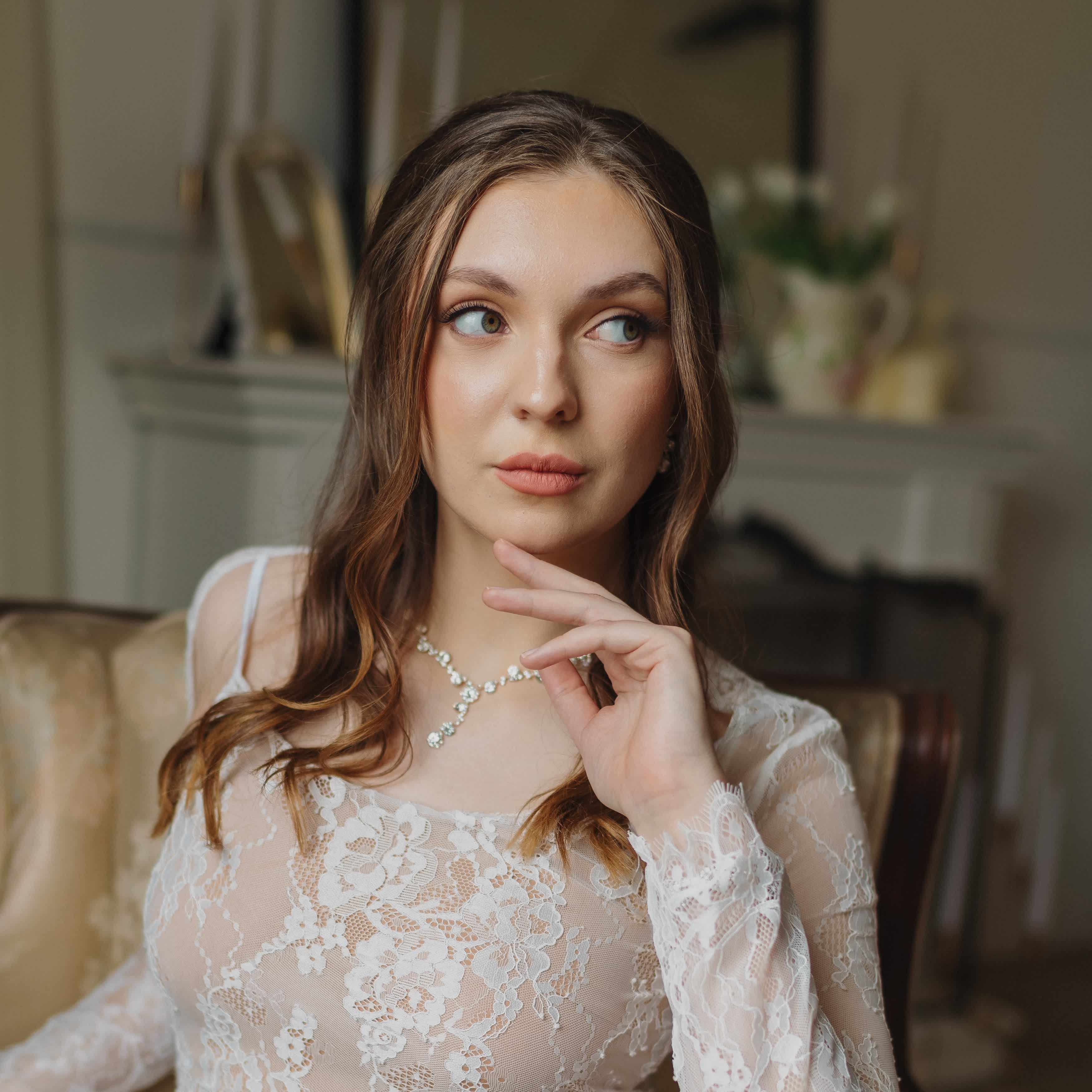 Bride in a lace gown with soft waves, resting her hand near her face.